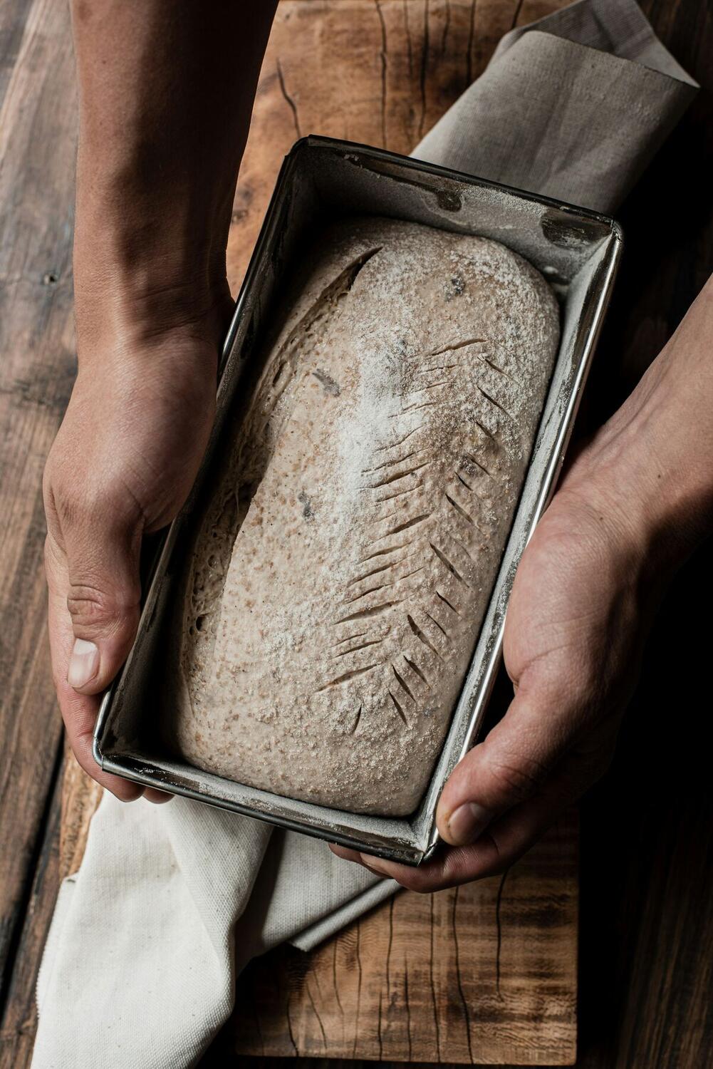 Two hands holding a loaf pan with a scored, unbaked sourdough loaf above a wooden table